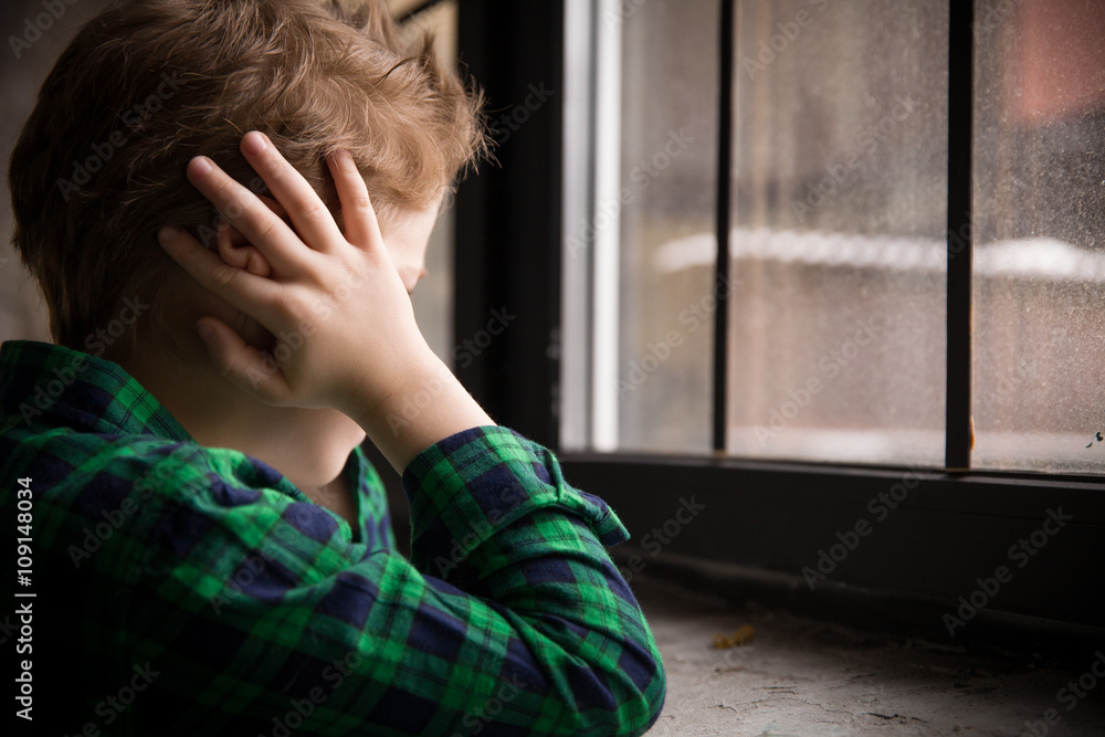 Little boy standing behind the window in sad mood. Sad Teenager looking ...
