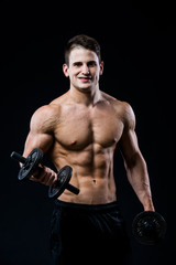 Wall Mural - Handsome power athletic man training pumping up muscles with dumbbells in a gym. Fitness muscular body isolated on black background. Looking to the camera.