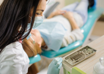 Wall Mural - Young beautiful woman dentist doing first check-up for female patient at the dental office. Doctor choosing dental tools, wearing mask and gloves. Medical equipment.