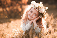 Smiling Baby Girl 3-4 Year Old Wearing Flower Wreath Outdoors. Sitting In Meadow. Looking At Camera. Childhood. 