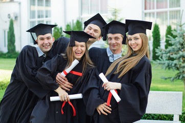 Graduates embrace, enjoy and look at the camera on the graduation ceremony. Happy graduation day. 5 graduates hold his graduate diplomas in their hands.