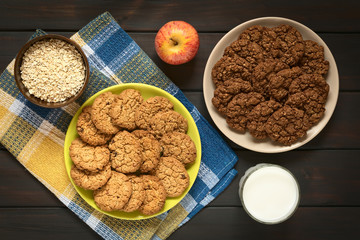 Wall Mural - Chocolate and apple oatmeal cookies on plates with an apple, a glass of milk and a bowl of raw oatmeal, photographed on dark wood with natural light