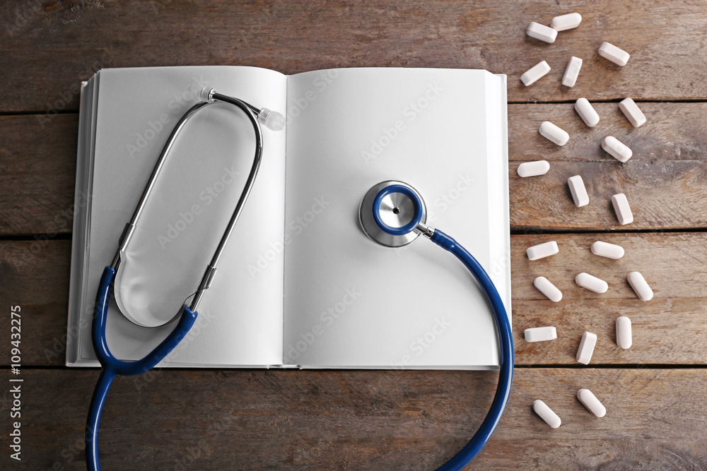 Stethoscope with open book and pills on wooden background. Medical literature concept