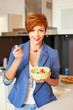 © guruXOX - Smiling young woman eating fresh fruits in modern kitchen
