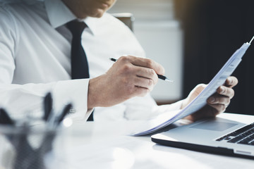 Close-up of professional businessman working at his office with documents, young lawyer using laptop computer and signing contract at comfortable interior, flare light