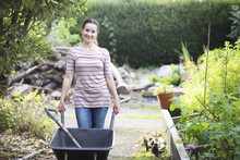 Portrait Of Female Gardener Pushing Wheelbarrow In Rustic Organic Garden