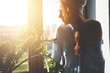© WHstudio Leushin N - Pretty hipster girl with bare shoulder sitting on the windowsill waiting for her friends to go out for a walk on sunny summer day with sunbeams and lens flare, enjoying urban view through the window