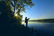 Silhouette Of A Fisherman With A Fishing Rod On The Shore Of The Lake, The River In The Morning. The Rays Of The Rising Sun Filtering Through The Tree Leaves
