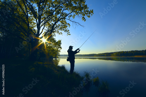 Fotografie, Obraz silhouette of a fisherman with a fishing rod on the shore of the lake, the river in the morning