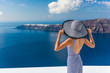 Europe summer vacation travel destination luxury living woman looking at view of Mediterranean Sea and Santorini island Oia village. Elegant tourist lady in fashion back dress and floppy sun hat.