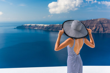 Fototapete - Europe summer vacation travel destination luxury living woman looking at view of Mediterranean Sea and Santorini island Oia village. Elegant tourist lady in fashion back dress and floppy sun hat.