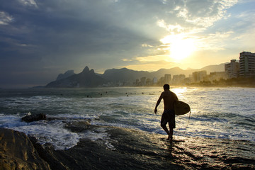 Surfer walking over rocks at sunset in Arpoador beach at Ipanema in Rio de Janeiro