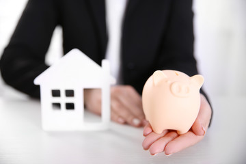 Wall Mural - Savings concept. Woman holding piggy bank in one hand and house figure in another, close up