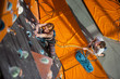 Strong woman climber with carbines and rope on an indoor rock-climbing wall is climbing up. Man standing on the ground insuring the climber