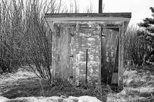 A Wooden Vintage Men And Women Outhouse With A Broken Door Sitting In Front Of Bare Trees In Black And White