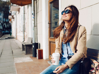 Beautiful and young girl in a coat and scarf and sunglasses sitting on the bench. Woman drinking coffee and laughing. Summer.