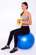 Staying fit. Young beautiful woman in sportswear with perfect bodie doing exercise with dumbbells and looking away with smile while sitting on fitness balls over white isolated background.
