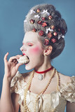 Young woman greedy eating marshmallow in light corset over blue background. Updo baroque hairstyle with dry roses.