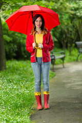 Wall Mural - Beautiful smiling young woman walking in a park on a rainy day. She is wearing red raincoat, rubber boots and holding a red umbrella.