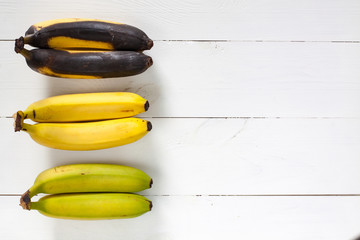 Green, yellow and black bananas arranged in a row on white wooden table.