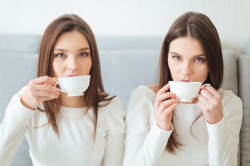 Two sisters twins sitting on sofa and drinking coffee