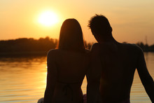 Young Couple Sitting On Lake Pier, Sunset Silhouette