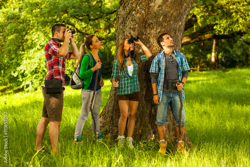 Plakat Grupa młodzi przyjaciele stoi lasowym drzewem na pięknym letnim dniu. Jedna kobieta używa lornetki.