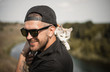 © endrews21 - a bearded man in a black baseball cap, black shirt and shorts playing with white fluffy cat on top of a cliff on the background of the river and the forest