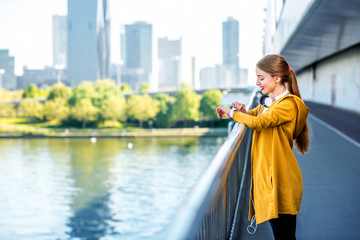 Young sport woman in yellow sweater looking at smart watch on the modern bridge with skyscrapers on the background. Morning exercise in megacity