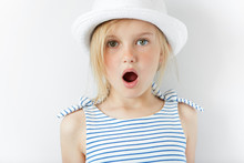 Close Up Portrait Of Amazed Adorable Little Girl In White Hat And Striped Dress, Having Fun Indoor, Looking At The Camera In Excitement, Astonished With Something. Human Face Expressions And Emotions