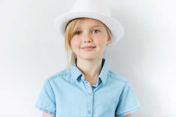 Close up portrait of adorable blonde little girl wearing white hat and blue shirt smiling and having fun indoor. Cute preschool female looking at the camera. Isolated shot of beautiful Caucasian child