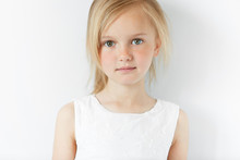 Close Up Shot Of Cute Fashionable Child With Green Eyes And Blonde Hair Weraing White Dress, Looking At The Camera With Serious Expression. Portrait Of Adorable Little Girl. Happy Childhood Concept