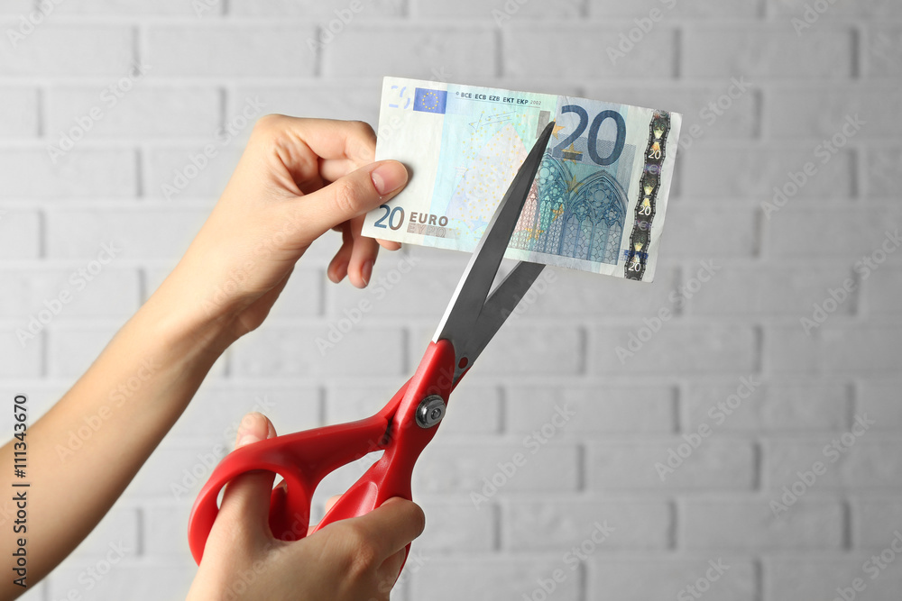 Hands with scissors cutting money on white brick wall background