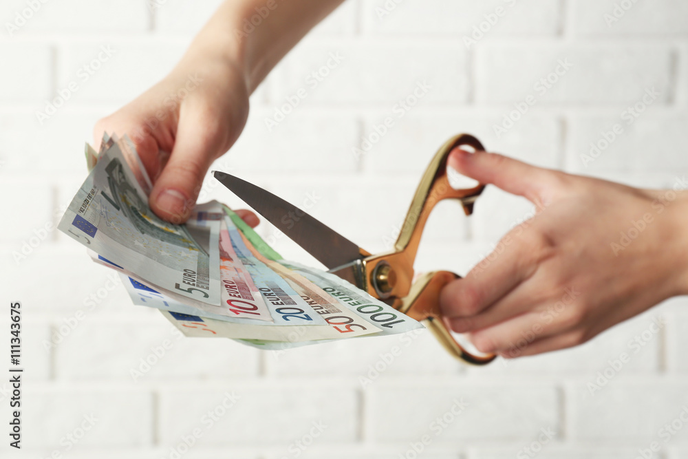 Hands with scissors cutting money on white brick wall background
