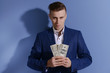 © Africa Studio - Attractive man in a suit holding fan of dollar banknotes on blue background