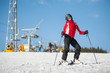 © anatoliy_gleb - Young female skier wearing helmet, red jacket and ski goggles standing with skis on mountain top at a winter resort in sunny day with ski lifts and blue sky in background.