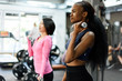 Side view of slim black african-american fitness instructor and asian lovely woman doing fitness exercise work with dumbbells together in the gym