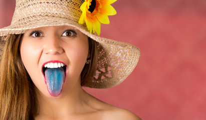 Pink background with a beautiful girl using a nice hat with a sunflower, girl sticking out her blue tongue