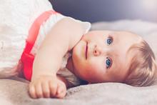 Portrait Of Cute Adorable Blonde Caucasian Smiling Baby Child Girl With Blue Eyes In White Dress With Red Bow Lying On Bed Looking Up Dreaming, Fairy Tale Sun Light Leak Ray Beam From Above Behind