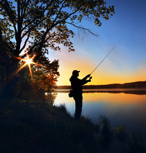 Silhouette Of A Fisherman With A Fishing Rod On The Shore Of The Lake, The River In The Morning. The Rays Of The Rising Sun Filtering Through The Tree Leaves
