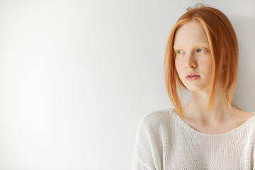 Isolated headshot of pretty teenage girl with ginger hair and perfect freckled skin holding arms folded. Cute young female wearing stylish white top, looking away at the blank copy space wall