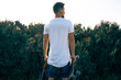 Young male with a skateboard in a blank white t-shirt is standing with his back to the camera. A man with a backpack is looking aside on a plant background.