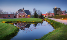 Landscape In Dutch Village With Beautiful House Reflected In Water Canal, Courtyard With Green Grass , Yellow Flowers And Road With Illumination At Dusk In Netherlands.
