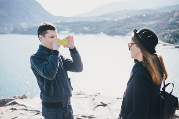 man taking picture of his girlfriend using his smartphone near seaside and mountains. travel togethe