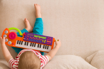 Top view on small cute toddler boy sitting on the sofa and playing on the toy piano. A little boy learning to play piano listening to music. Early development for toddler. Musical education for kids