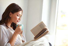 Young Woman At Home Sitting Near Window Relaxing In Her Living Room Reading Book And Drinking Coffee Or Tea