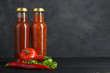 red sauce in a glass bottle near the red hot pepper and greens leaves on black wooden background