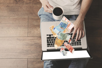 Wall Mural - Financial concept. Make money on the Internet. Young man sitting on floor with laptop and cup of coffee in room