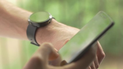 Man hands gestures on a round screen modern smartwatch and a smartphone device. Connecting them togheter to be sync. Beautiful green exterior outdoor nature background bokeh. 
