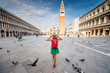 Young female traveler with hat and backpack standing on San Marco square with tower and basilica on the background in Venice. Back view with copy space